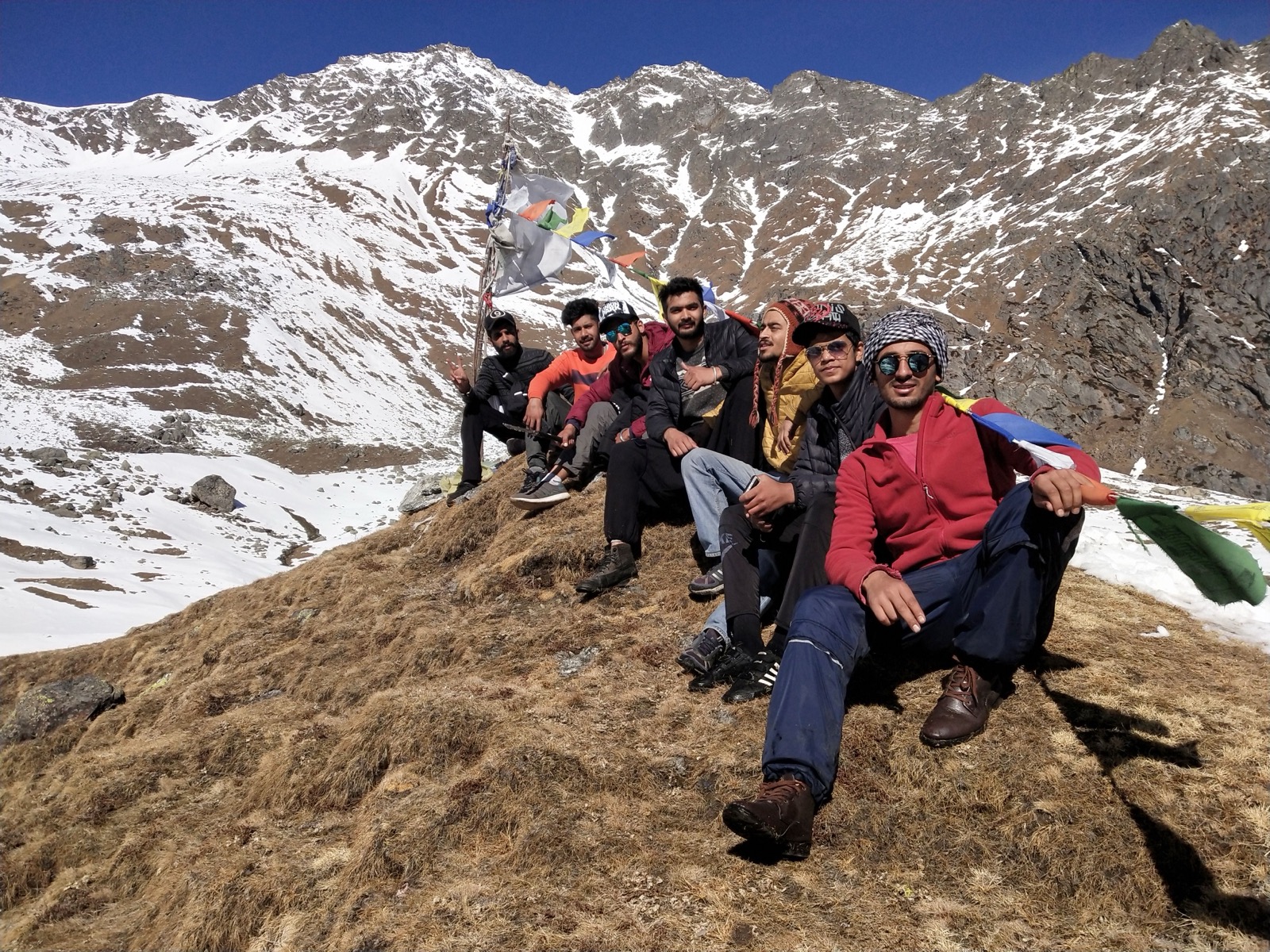 Trekkers resting at Yulla Kanda summit with prayer flags and snow-covered Himalayan peaks